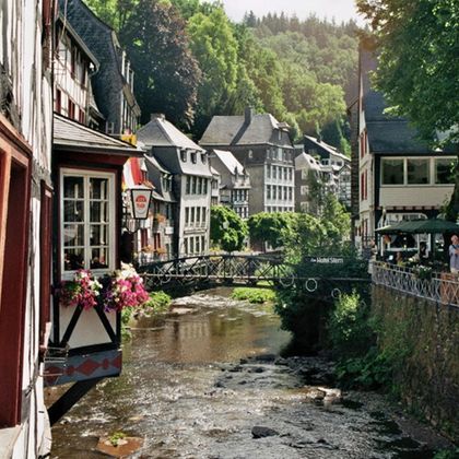 Eine malerische Stadtansicht mit traditionellen Gebäuden am Fluss. Grüne Hügel umgeben die Szene und verleihen ihr einen idyllischen Charakter.