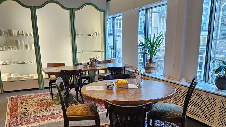 Ein gemütlicher Raum mit Holzstühlen und einem runden Tisch. Große Fenster bringen viel Licht und eine Pflanze sorgt für eine frische Atmosphäre.