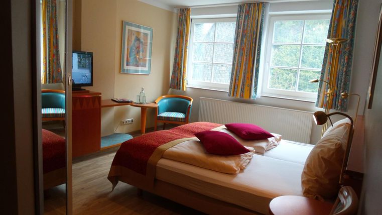 Ein helles Schlafzimmer mit einem Doppelbett und bunten Kissen. Es gibt einen Fernseher und zwei bequeme Stühle vor einem Fenster.