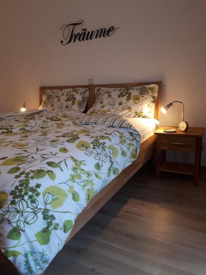 Ein gemütliches Schlafzimmer mit einem Doppelbett und einer natürlichen, blühenden Bettwäsche. An der Wand steht das Wort "Träume" und es gibt zwei Nachttischlampen.