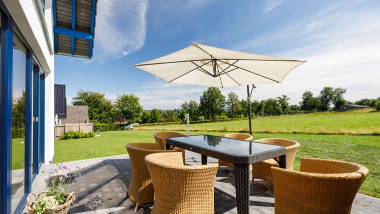 Ein sonniger Essbereich im Freien mit einem Tisch und Stühlen. Im Hintergrund ist eine grüne Wiese und ein klarer Himmel zu sehen.