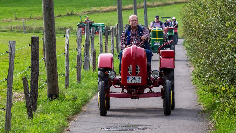 Ein roter Traktor fährt auf einem schmalen Weg, umgeben von grünen Wiesen. Im Hintergrund sind weitere Traktoren zu sehen.