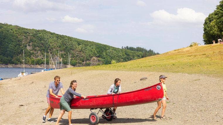 Eine Gruppe von vier Personen zieht ein rotes Kanu an den Strand. Im Hintergrund sind grüne Hügel und ein ruhiger Wasserbereich zu sehen.