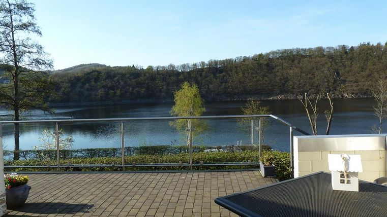 Ein ruhiger See mit grünen Ufern und sanften Hügeln im Hintergrund. Auf der Terrasse im Vordergrund stehen Pflanzen und ein Tisch.