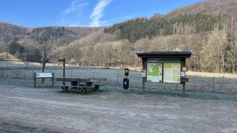 Ein ruhiger Ort mit einem Informationsstand und einer Bank in der Natur. Im Hintergrund sind sanfte Hügel und ein blauer Himmel zu sehen.