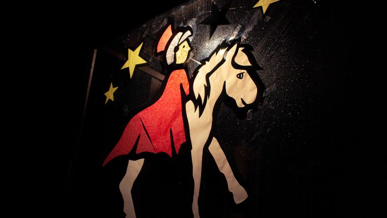 Ein farbenfrohes Papierbild zeigt eine Figur in Rot, die auf einem weißen Pferd reitet. Im Hintergrund sind einige gelbe Sterne.