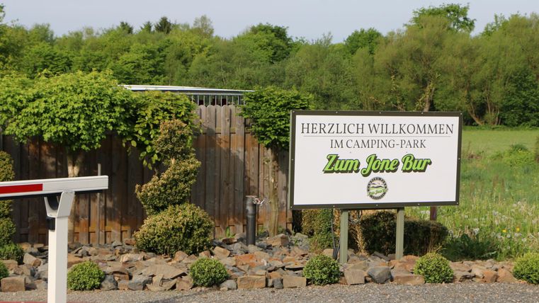 Ein Willkommensschild vor dem Camping-Park "Zum Jonen Bur". Die Umgebung ist grün und mit Bäumen und Sträuchern bewachsen.