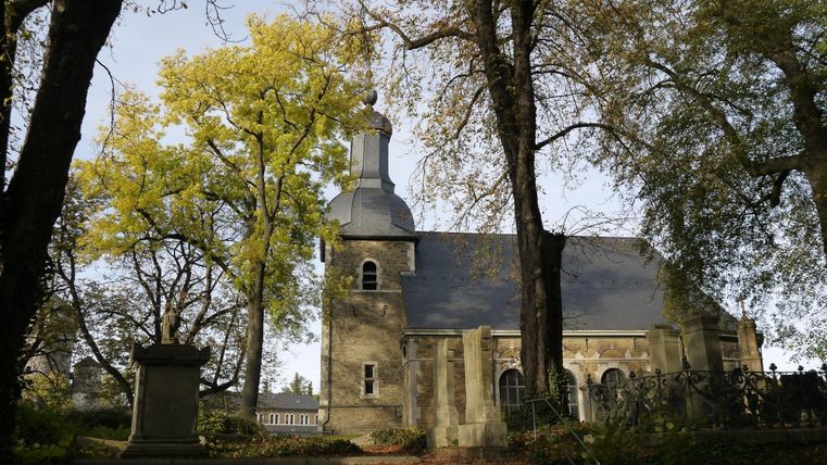 Eine alte Kirche umgeben von großen Bäumen. Der Herbst zeigt sich mit bunten Blättern und sanftem Licht.