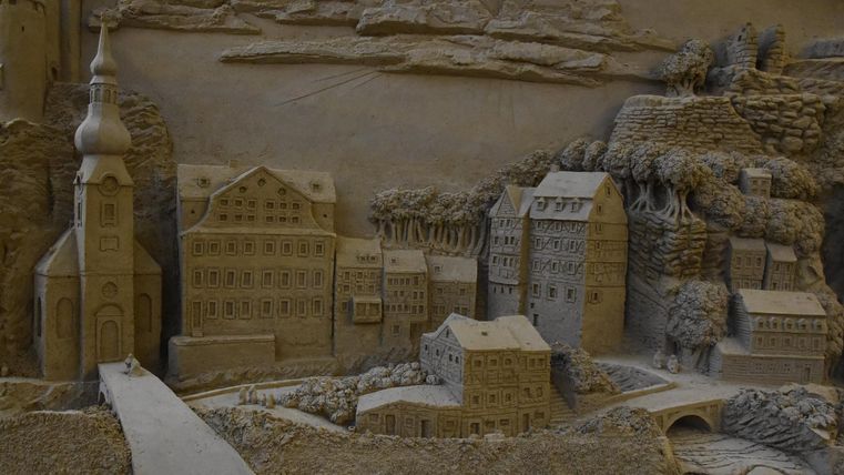 Eine detaillierte Sandkunst-Darstellung einer Stadt mit Gebäuden, einer Brücke und einer Kirche. Die Landschaft zeigt natürliche Felsen im Hintergrund.