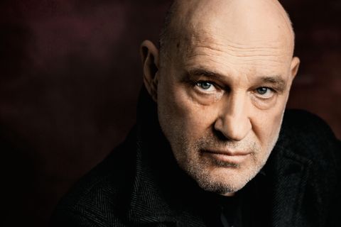 Ein älterer Mann mit Glatze und ernstem Gesichtsausdruck schaut direkt in die Kamera. Der Hintergrund ist dunkel und unscharf, was den Fokus auf sein Gesicht lenkt.