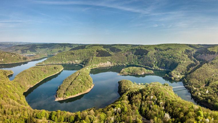 Eine panoramische Sicht auf einen malerischen Fluss mit sanften Kurven, umgeben von grünen Hügeln. Der klare Himmel und die ruhige Wasseroberfläche schaffen eine friedliche Atmosphäre.
