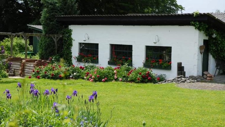Ein charmantes, weißes Haus mit bunten Blumen am Fenster. Der Garten ist üppig grün, mit vielen blühenden Pflanzen.