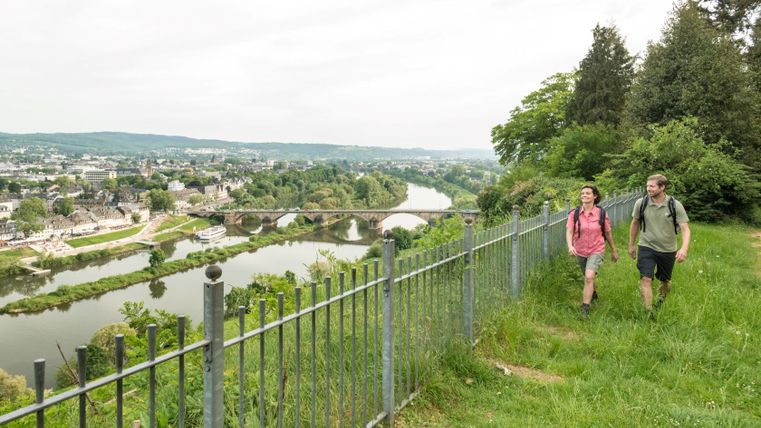 Zwei Wanderer gehen entlang eines Zauns mit Blick auf die Stadt Trier und die Mosel.