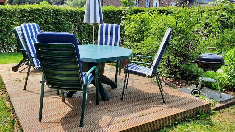 Ein einladender Gartenbereich mit einem runden Tisch und Stühlen auf einer Holzterrasse. Im Hintergrund befindet sich ein Grill und üppiges Grün.