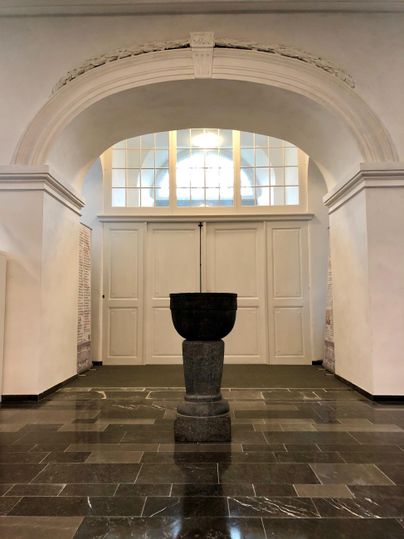 Ein schöner Innenraum mit hohen Decken und großen Fenstern. In der Mitte steht ein elegantes Steingefäß auf einem Sockel.