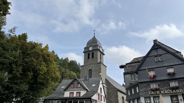 Ein malerischer Platz mit historischen Gebäuden und einer auffälligen Kirchturm. Der Himmel ist teilweise bewölkt, und es gibt Bäume im Hintergrund.