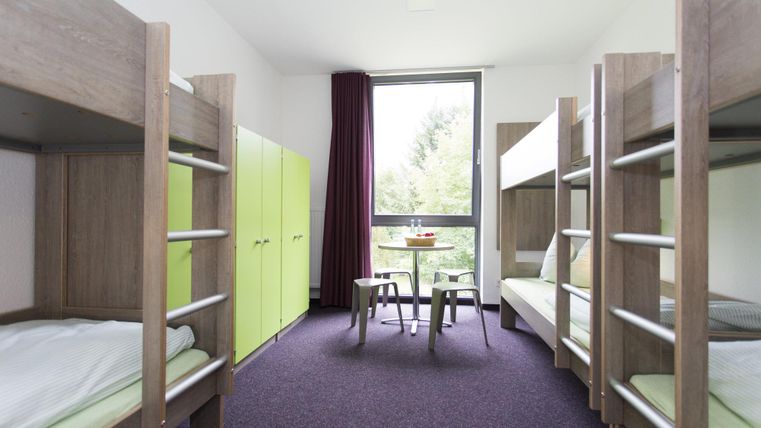 Ein modernes Zimmer mit zwei Etagenbetten und einem kleinen Tisch. Große Fenster lassen viel Licht herein und es gibt grüne Schränke.