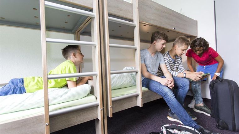 Eine Gruppe von vier Jugendlichen in einem Schlafsaal. Zwei sitzen auf einem unteren Bett, während ein anderer auf dem Bett liegt und eine Person etwas zeigt.