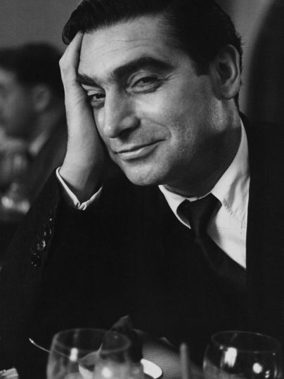 Ein lächelnder Mann sitzt an einem Tisch, mit einer Hand an seinem Kopf. Im Hintergrund sind Gläser und eine gedämpfte Atmosphäre sichtbar.