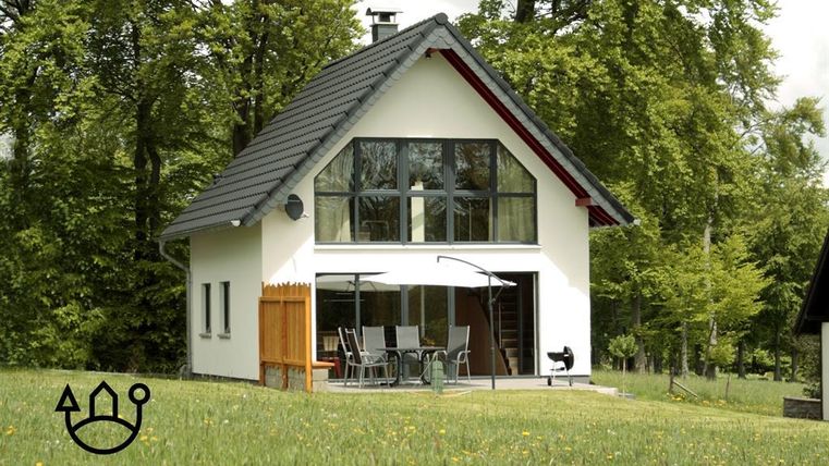 Ein modernes Ferienhaus umgeben von grüner Wiese und hohen Bäumen. Es bietet eine helle Fassade mit großen Fenstern und einer einladenden Terrasse.
