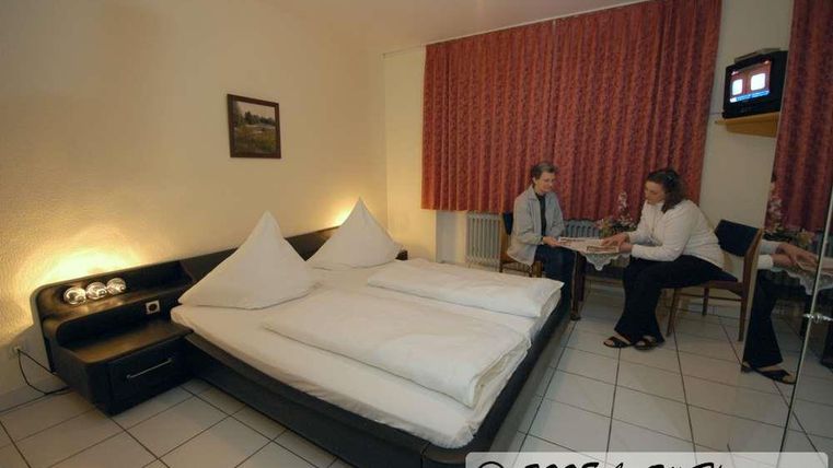 Ein einfaches Hotelzimmer mit einem Doppelbett und zwei Personen, die am Tisch sitzen. Die Wände sind mit roten Vorhängen dekoriert, und es gibt einen Fernseher im Raum.