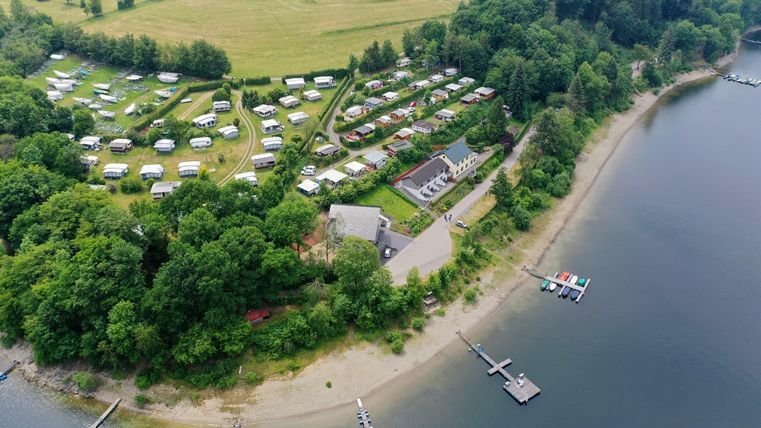 Ein schöner Campingplatz am Ufer eines Gewässers, umgeben von Bäumen. Mehrere Zelte sind auf dem Gelände verteilt, und es gibt Stege für Boote.