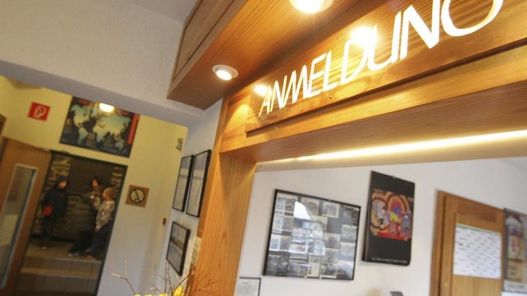 Ein Empfangsbereich mit einer Holztheke und der Aufschrift "ANMELDUNG". Im Hintergrund sind Gäste zu sehen und einige Wandbilder.