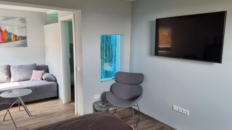 Ein modernes Zimmer mit einem großen Fenster und einem Fernseher an der Wand. Es gibt ein bequemes Sofa und einen Stuhl in der Ecke.