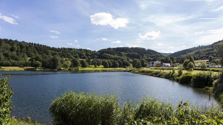 Ein ruhiger See umgeben von grünen Wiesen und sanften Hügeln. Der Himmel ist blau und mit einigen weißen Wolken versehen.