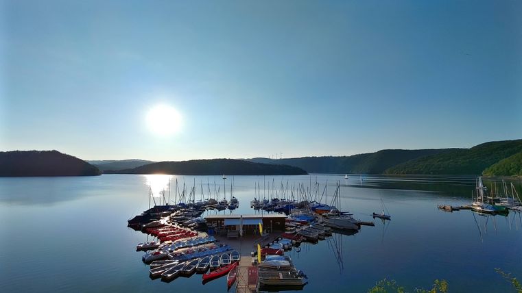Ein ruhiger Hafen mit vielen Booten auf einem stillen See. Die Sonne scheint über der Landschaft und wirft sanfte Reflexionen auf das Wasser.