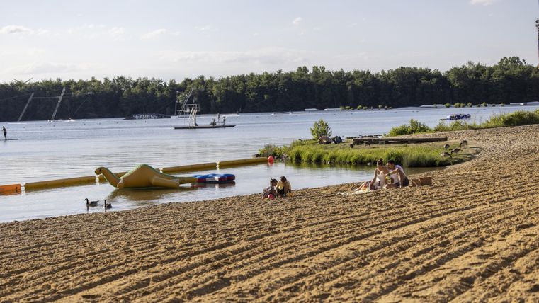 Ein Strand mit feinem Sand und ruhigem Wasser. Im Hintergrund sind einige Menschen und ein Boot zu sehen.