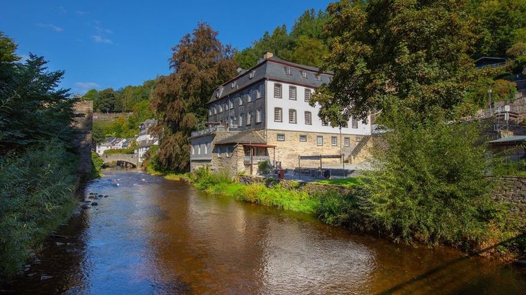 Ein malerisches Gebäude neben einem ruhigen Fluss, umgeben von Bäumen und Hügeln. Der Himmel ist klar und blau.
