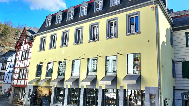 Ein modernes, gelbes Gebäude mit mehreren Fenstern und markanten Jalousien. Die Architektur steht im Kontrast zu den traditionellen Fachwerkhäusern in der Umgebung.