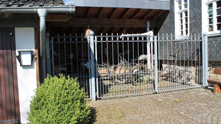 Ein Eingang mit einem metallenen Tor und einem Sichtschutz. Im Hintergrund sind Holzreste und ein Gebäude sichtbar.