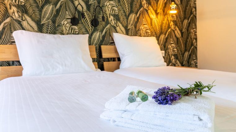 Ein gemütliches Schlafzimmer mit zwei Betten und einer eleganten Pflanzen-Tapete. Frisch gefaltete Handtücher und ein kleiner Strauß Lavendel liegen dekorativ auf dem Bett.