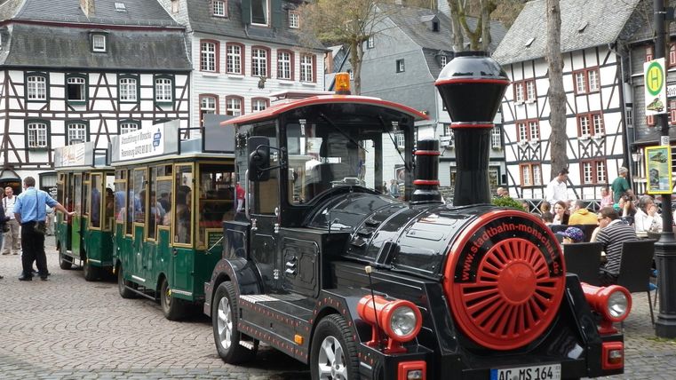 Eine kleine Bahn aus einem schwarzen Lokomotiv mit roten Akzenten parkt in einer malerischen Stadt mit Fachwerkhäusern. Die Straße ist gepflastert und es sind einige Personen in der Nähe zu sehen.
