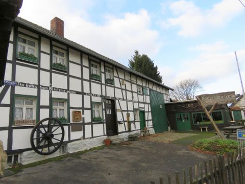 Ein traditionelles Fachwerkhaus mit weißen Wänden und dunklen Balken. Im Vordergrund steht ein großer Wagenrad und der Garten ist gepflegt.