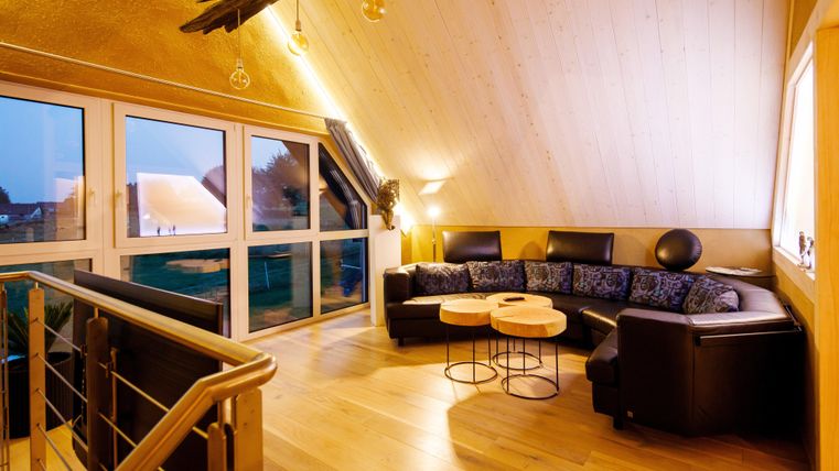 Ein gemütlicher Wohnraum mit einem großen, dunklen Sofa und zwei kleinen Tischen. Große Fenster bieten Blick auf die Umgebung und sorgen für viel natürliche Helligkeit.