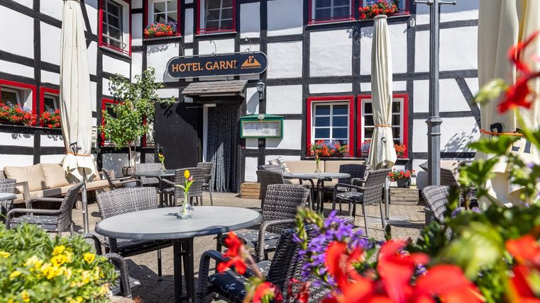 Eine gemütliche Terrasse vor einem Hotel mit schönen Pflanzen und Blumen. Es gibt Tische und Stühle, die zum Entspannen einladen.