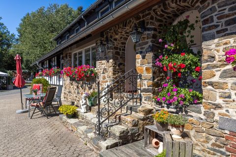 Ein einladendes Haus mit Steinfassade und bunten Blumenampeln. Vor dem Haus stehen Stühle und ein Sonnenschirm.