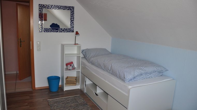 Ein gemütliches Schlafzimmer mit einem Einzelbett und einer hellen Wand. Im Raum befindet sich ein Regal und ein blauer Mülleimer.