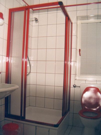 Ein Badezimmer mit einer Duschkabine und weißen Fliesen. Auf der linken Seite steht ein Waschbecken, und rechts befindet sich eine Toilette.
