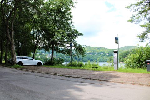 Eine ruhige Straße mit Bäumen und einem weißen Auto. Im Hintergrund ist ein See und hügelige Landschaft zu sehen.