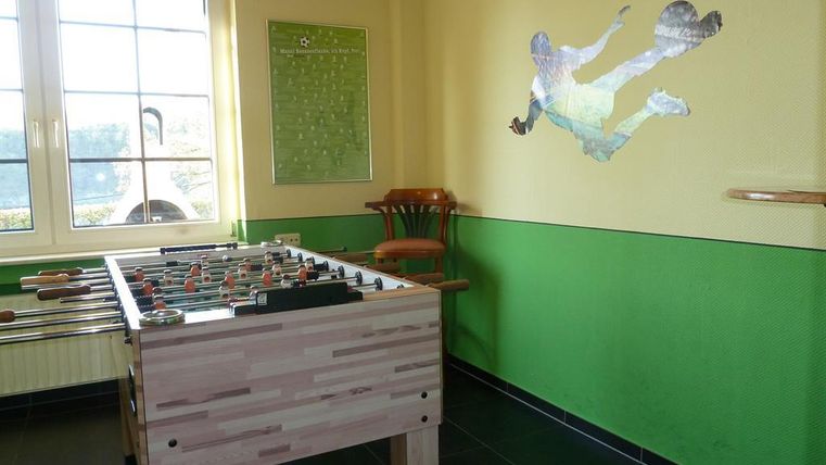 Ein Spielebereich mit einem Kicker und einer bunten Wandgestaltung. Ein Stuhl steht in der Ecke, und ein großes Fenster bietet Tageslicht.