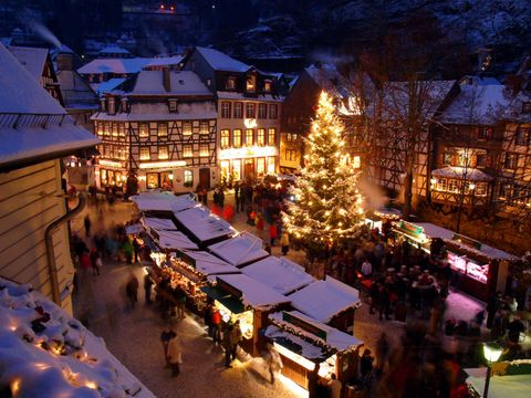 Ein schöner Weihnachtsmarkt mit festlich beleuchteten Ständen und einem großen Weihnachtsbaum. Die winterliche Atmosphäre wird durch den Schnee und die beleuchteten Gebäude verstärkt.