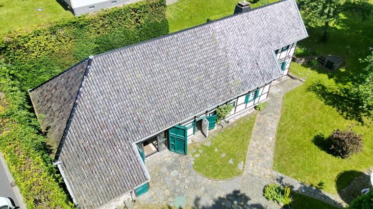 Ein traditionelles Haus mit einem schrägen Dach, umgeben von grünem Rasen und Bäumen. Der gepflasterte Weg führt zur Haustür.