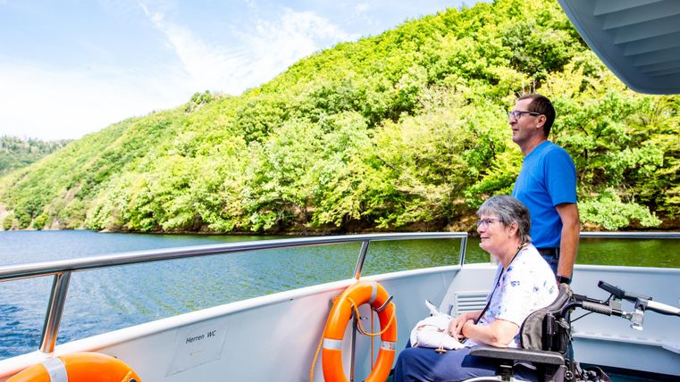 Eine Person im Rollstuhl genießt die Aussicht auf den See, während eine andere Person daneben steht. Grüne Hügel und ein klarer Himmel sind im Hintergrund zu sehen.