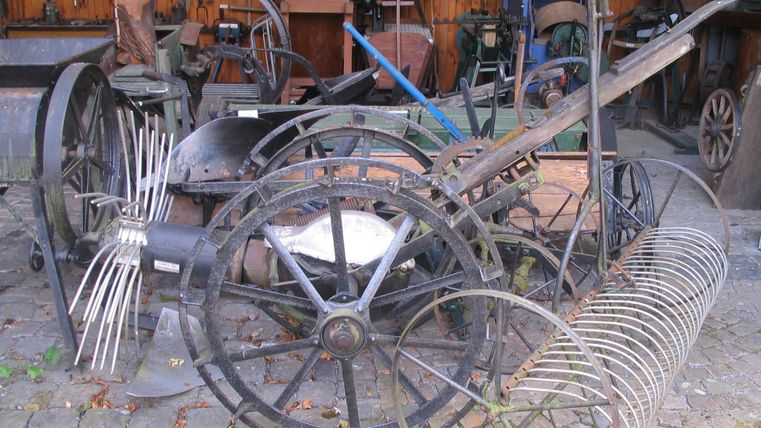 Eine Ansammlung von alten landwirtschaftlichen Geräten und Maschinen. Die Objekte sind aus Metall und Holz und liegen in einer Scheune.