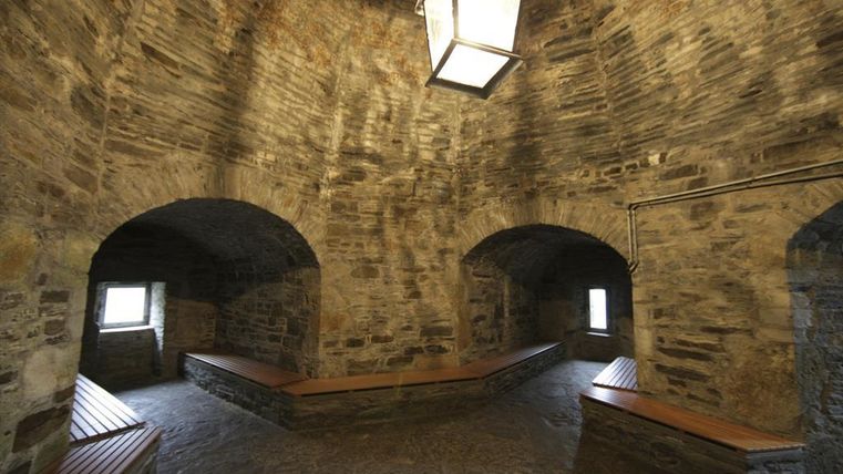 Ein historischer Raum mit steinernen Wänden und Bänken aus Holz. Eine Deckenlampe sorgt für sanftes Licht und beleuchtet die Atmosphäre.