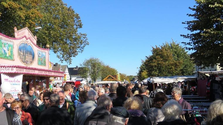 Ein belebter Markt mit vielen Menschen. Bunte Stände und herbstliche Bäume sind sichtbar.
